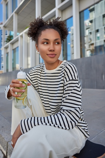 Vertical shot of pretty thoughtful millennial girl looks away drinks refreshing beverage after riding skateboard dressed in striped jumper and trousers poses outside