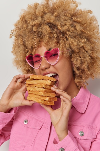 Vertical shot of pretty young woman with curly bushy hair bites big pile of waffles wears heart shaped sunglasses and jacket has sweet tooth poses indoor Delicious appetizing homemade dessert