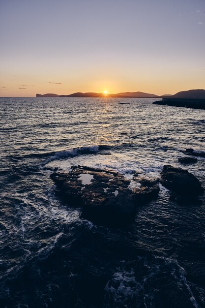 遠くの山の後ろに太陽が輝いている海の岩の垂直方向のショット