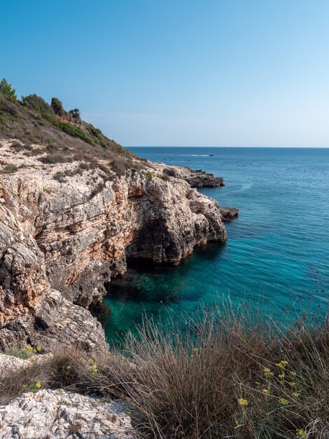 Vertical shot of a rocky cliff with a blue-green sea on a clear sky background