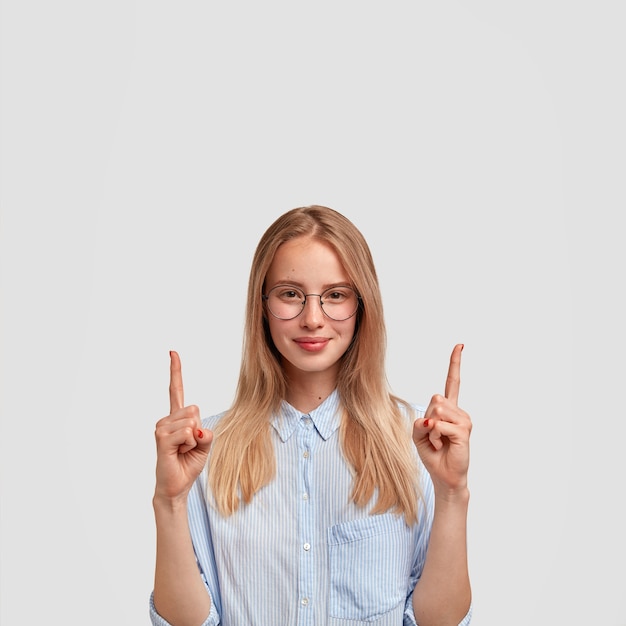 Vertical shot of satisfied young European female with pleased expression, points upwards with index fingers, dressed in fashionable shirt, shows something above, isolated over white wall.