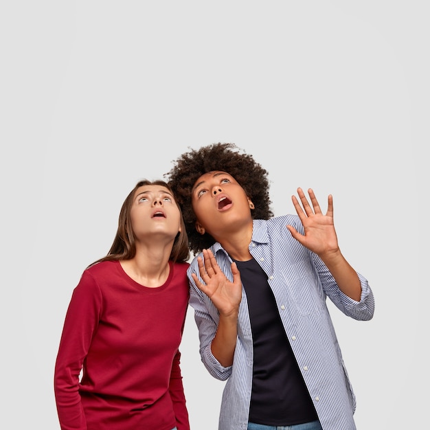 Vertical shot of scared best friend raise heads, have stupefied look at ceiling, afraid of heavy thing falling down, keep hands in protective gesture, dressed in fashionable clothes, isolated on white