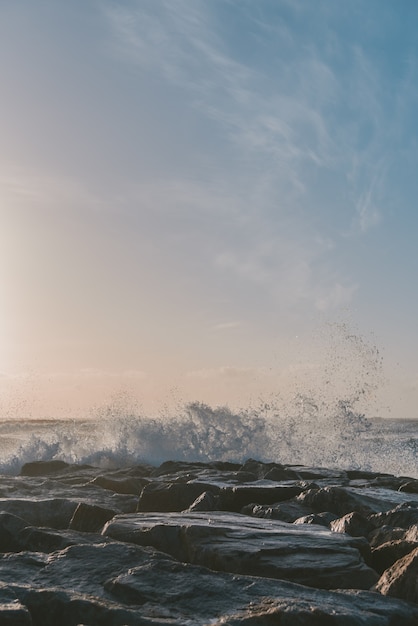 Vertical shot of the sea waves hitting the rocks under a blue sky