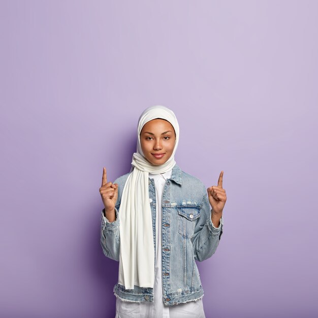 Vertical shot of self confident muslim woman with dark skin, points to top, shows great copy space upwards for customers, wears white silk scarf and denim coat, isolated on purple wall