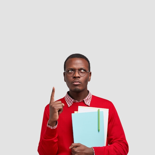 Vertical shot of serious black man has thoughtful expression, dressed in red sweater, points with index finger on ceiling