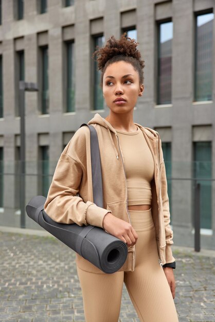 Vertical shot of serious curly haired woman dressed in brown sportswear carries rolled karemat goes in for sport regularly looks away poses against modern building in city Healthy lifestyle concept