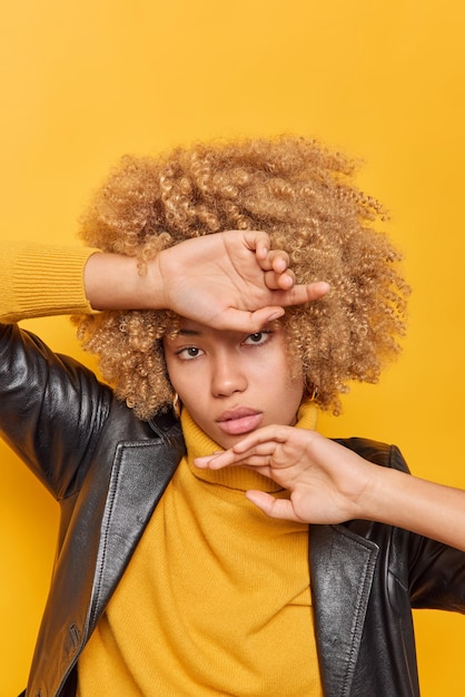 Free photo vertical shot of serious woman keeps hands near face touches jawline has curly bushy hair dressd in casual clothes looks self confident at camera isolated on yellow background. human face expressions