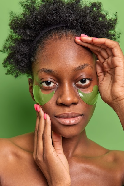 Vertical shot of serious young woman with natural curly hair applies hydrogel patches under eyes to reduce fine lines and puffiness stands shirtless against vivid green wall