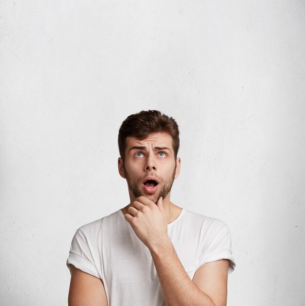 Vertical shot of shocked bearded man keeps mouth widely opened, looks in bewilderment upwards, noticies something unexpected, poses against white concrete wall