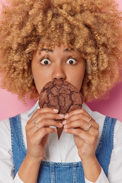 Free photo vertical shot of shocked curly haired young woman covers mouth with yummy chocolate cookie eats sweet food stunned to break diet dressed in casual clothes poses indoor unhealthy food concept