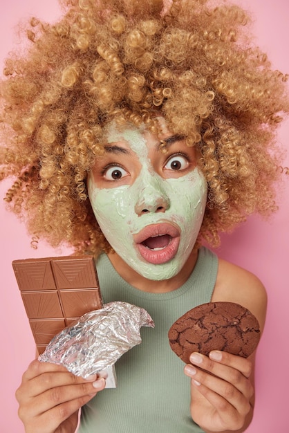 곱슬곱슬한 머리를 한 충격을 받은 유럽 여성의 세로 사진은 입을 크게 벌리고 분홍색 배경 위에 격리된 초콜릿과 달콤한 쿠키를 들고 있는 뷰티 클레이 마스크를 적용합니다.