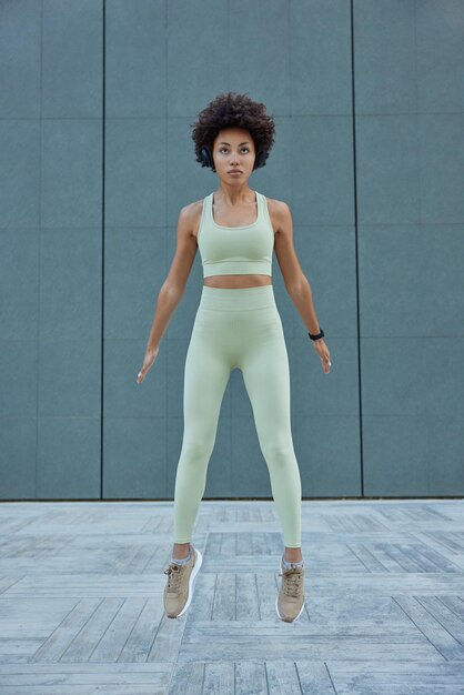 빈 회색 벽에 대고 날씬한 곱슬머리 여성 점퍼의 세로 샷은 자른 탑 레깅스를 입고 운동화는 운동복을 입고 규칙적인 운동을 합니다.