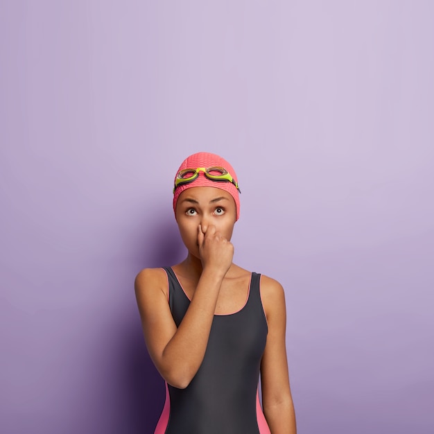 Vertical shot of slim woman holds breath, covers nose, preapres for diving under water, looks above