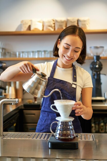 Vertical shot of smiling asian bartender barista in blue apron pouring water with small kettle brewi