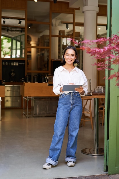 Vertical shot of smiling asian businesswoman waitress standing in front of cafe entrance inviting gu