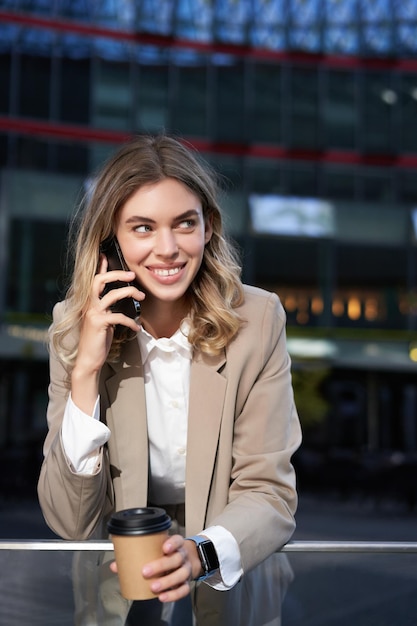 Free photo vertical shot of smiling businesswoman answer mobile phone call talking on smartphone and drinking c