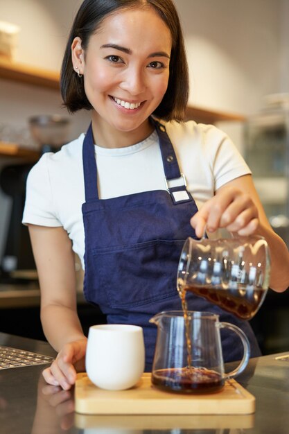 Вертикальный снимок улыбающейся милой девушки-бариста, наливающей фильтрованный кофе, делая заказ для клиента кафе