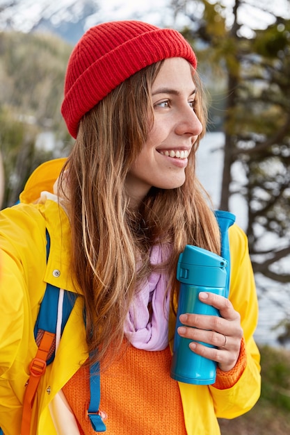 Vertical shot of smiling female traveler wears red hat, yellow coat, stretches hand, makes selfie with unrecognizable device