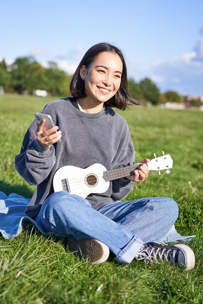Vertical shot of smiling korean girl sitting in park with ukulele and smartphone using mobile app to