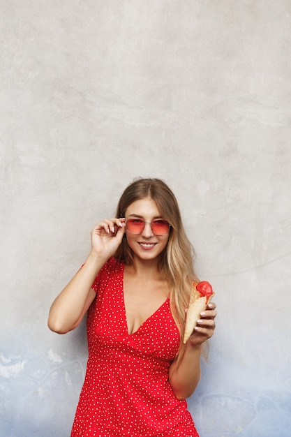 Вертикальный снимок улыбающейся женщины в солнцезащитных очках, наслаждающейся поездкой на летний райский остров и едящей сладкое мороженое