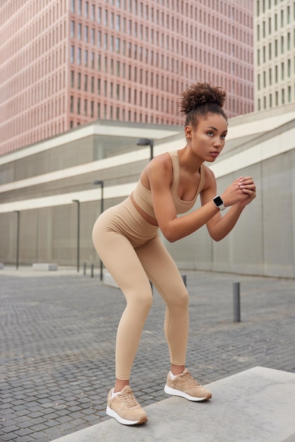 スポーティな若い女性の垂直ショットは、ベージュのスポーツウェアに身を包んだスクワットを行い、スニーカーのエクササイズは、都会の背景に対して屋外で定期的にポーズをとる人々のトレーニング健康的なライフスタイルの概念