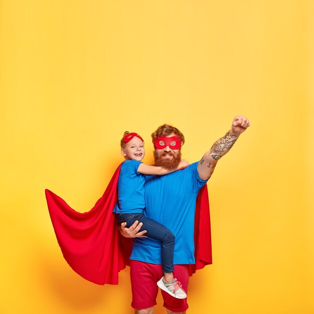 Vertical shot of strong red haired man in superhero costume, raises fist and makes flying gesture