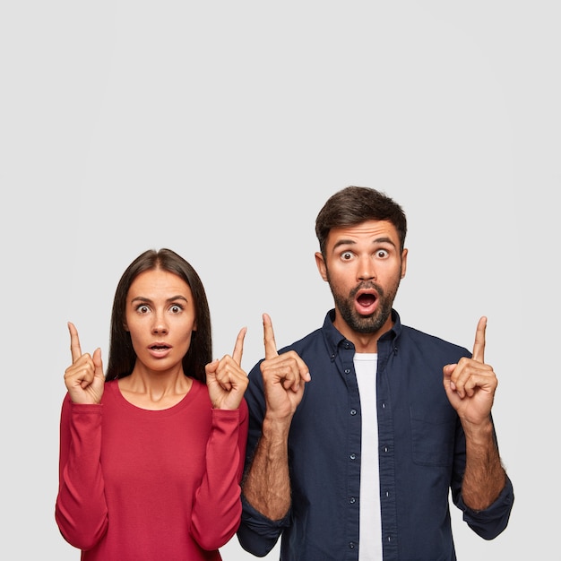 Vertical shot of stunned coworkers point with index fingers upwards, show free space for your advertisement, have eyes full of disbelief