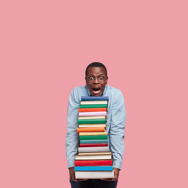Vertical shot of stupefied black man leans head on pile of books, wears round glasses, surprised with many tasks for seminar