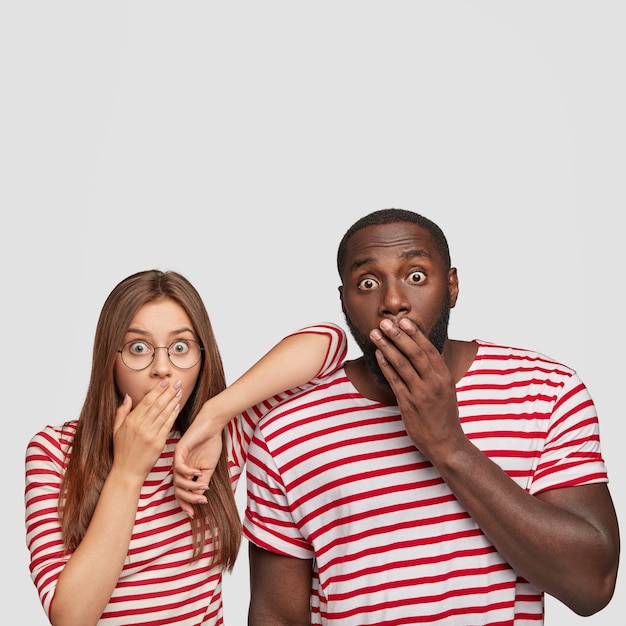 Vertical shot of stupefied dark skinned man with terrified expression, covers mouth with hand, dressed in striped clothes