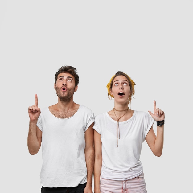 Vertical shot of stupefied female and male point both index fingers up, indicate something shocking, drop jaw, concentrated on ceiling