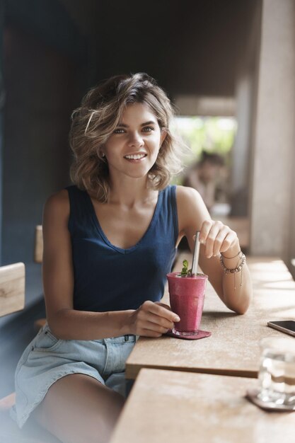 Vertical shot stylish female student take break drinking smoothie sit window bar indoor cafe smiling delighted camera talking have pleasant carefree conversation after university lectures
