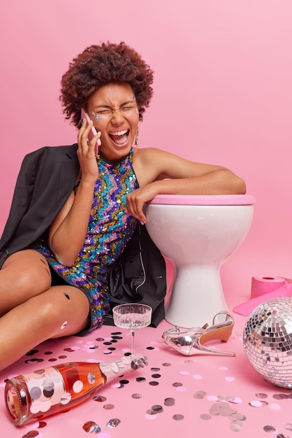 Vertical shot of stylish lady yells loudly has telephone conversation leans on toilet bowl poses in washroom after party with bottle of champagne disco ball and confetti against pink background