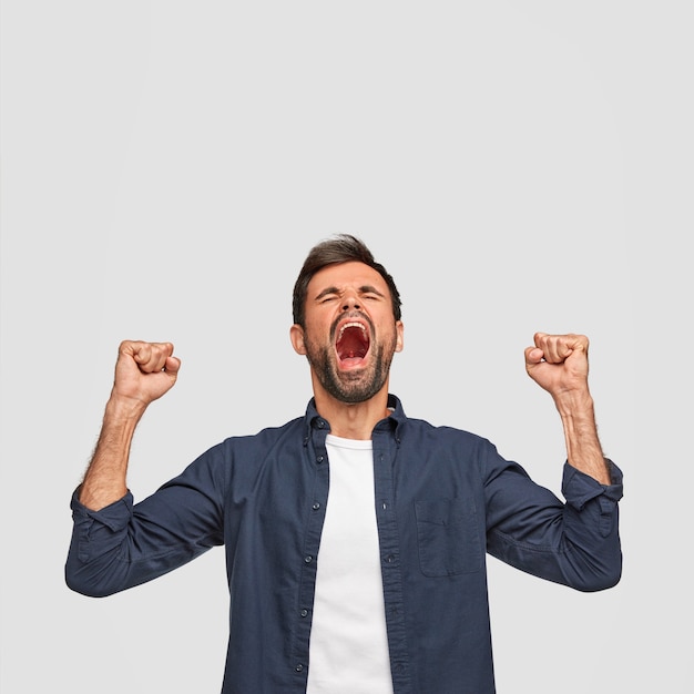 Vertical shot of successful male has overjoyed expression, clenches fists and opens mouth widely, exclaims with happiness, dressed casually, isolated over white wall with copy space upwards