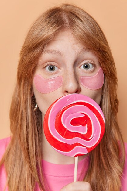 Vertical shot of surprised redhead girl has freckled skin applies hydrogel patches under eyes for skin treatment covers mouth with round sweet lollipop stands indoor Female teenager with candy