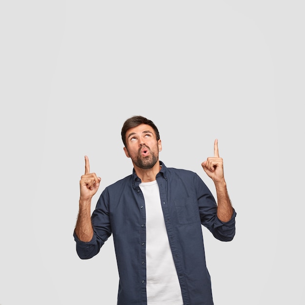 Vertical shot of surprised unshaven guy points with both index fingers upwards