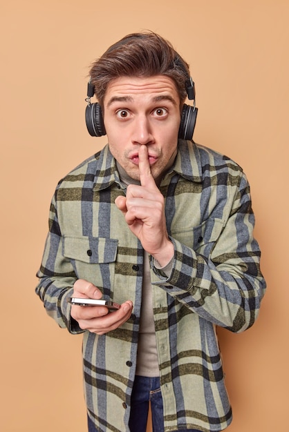 Free photo vertical shot of surprised young handsome man makes silence gesture spreads gossips holds mobile phone listens audio track via headphones dressed in checkered shirt isolated over brown background