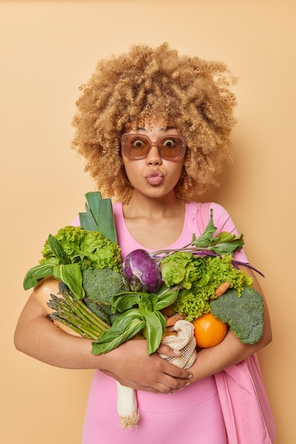 Вертикальный снимок удивленной молодой женщины, держащей губы округлыми, несет разнообразные продукты из свежих овощей, носит солнцезащитные очки, одетые в розовую спортивную одежду, ест здоровую пищу, изолированную на бежевом фоне