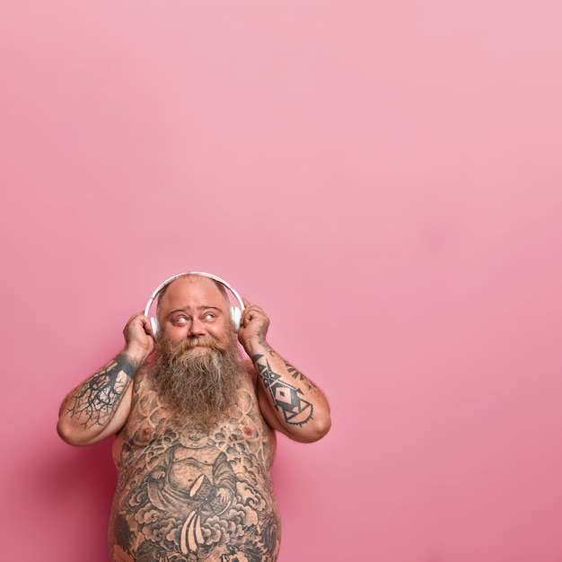 Vertical shot of thoughtful bearded man looks pleased above, keeps hands on headphones, thinks about something pleasant while listens music, has problem of obesity, tattooed naked body, stout tummy