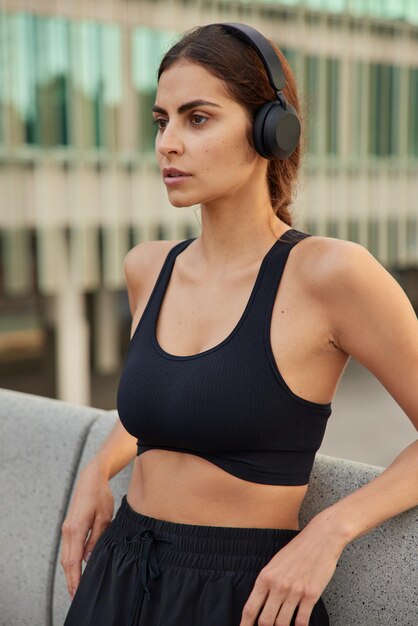 Vertical shot of thoughtful determined sportswoman dressed in sportswear rests after cardio training poses against blurred background listens motivation music via headphones has athletic healthy body