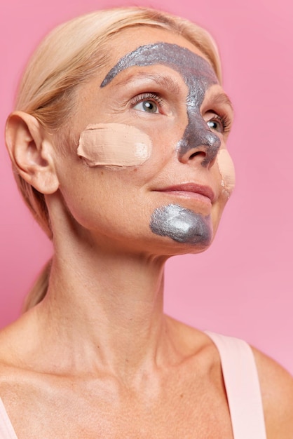 Вертикальный снимок вдумчивой женщины средних лет, которая наносит маску для лица, а тональный крем заботится о коже лица, сосредоточенной вдалеке, а светлые причесанные волосы изолированы на розовом фоне. Понятие красоты