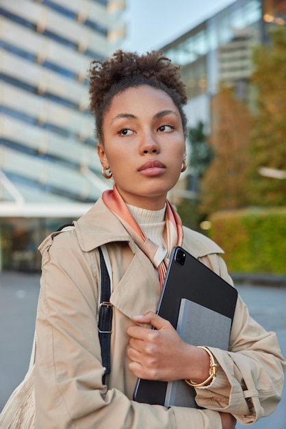 Vertical shot of thoughtful woman dressed in outerwear holds modern tablet and notebook focused away walks outside