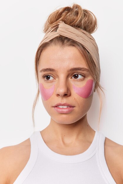 Vertical shot of thoughtful young European woman concentrated somewhere wears headband applies pink beauty pads under eyes to reduce puffiness dressed in t shirt isolated over white background