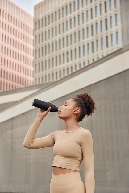 Vertical shot of tired curly sportswoman takes break after intensive training in urban setting drinks refreshing water poses outdoors wears beige tracksuit. People and healthy lifestyle concept