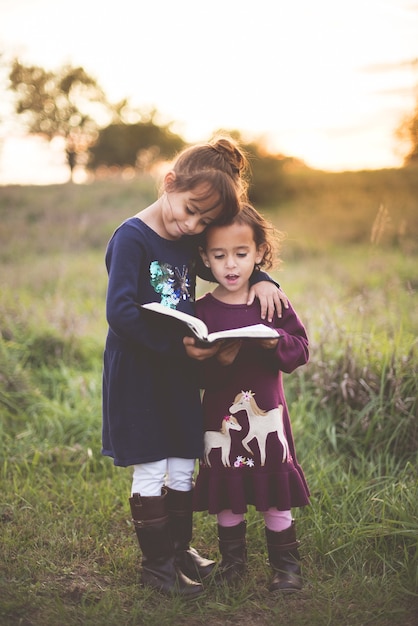 Vertical shot of two adorable little sisters reading the Bible at the park