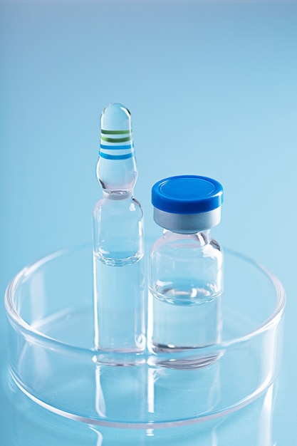 Vertical shot of two different glass vials with clear liquid in a glass dish on blue surface