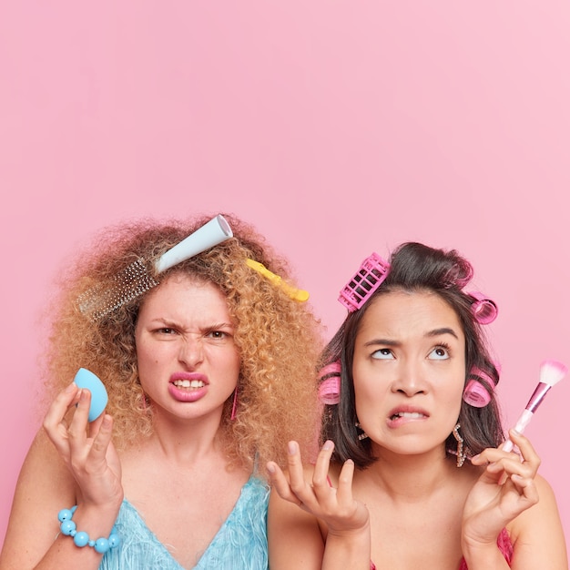화가 난 두 여성이 서둘러 파티 준비를 하는 세로 사진은 화장 도구를 사용하여 화장을 하고 웃는 얼굴은 위의 복사 공간이 있는 분홍색 벽에 함께 머리를 빗고 포즈를 취합니다.