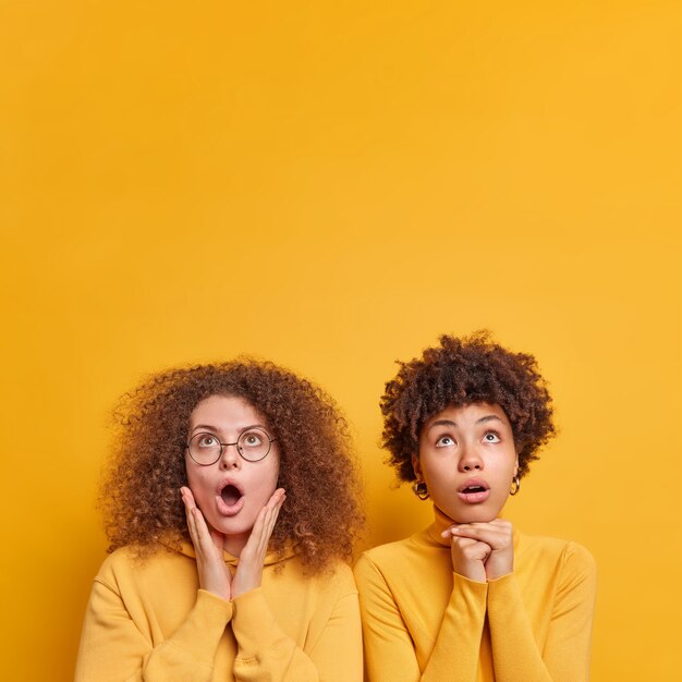위의 충격으로 응시하는 두 명의 다양한 여성의 세로 샷은 승진을 위해 노란색 벽 복사 공간 위에 자연스럽게 격리 된 옷을 입고 서로 밀접하게 입을 열어 유지합니다.