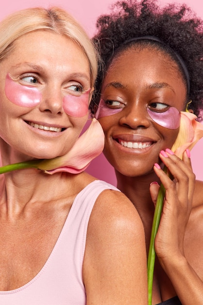 Vertical shot of two happy women take care of complexion apply beauty moisturising patches under eyes hold flower near face undergo skin care procedures smile gently pose against pink