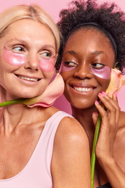 Вертикальный снимок двух счастливых женщин, которые заботятся о цвете лица, наносят косметические увлажняющие патчи под глаза, держат цветок рядом с лицом, проходят процедуры по уходу за кожей, мягко позируют на фоне розового.