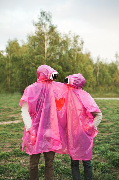 Vertical shot of two people looking at each other in VR headsets sharing a pink plastic raincoat